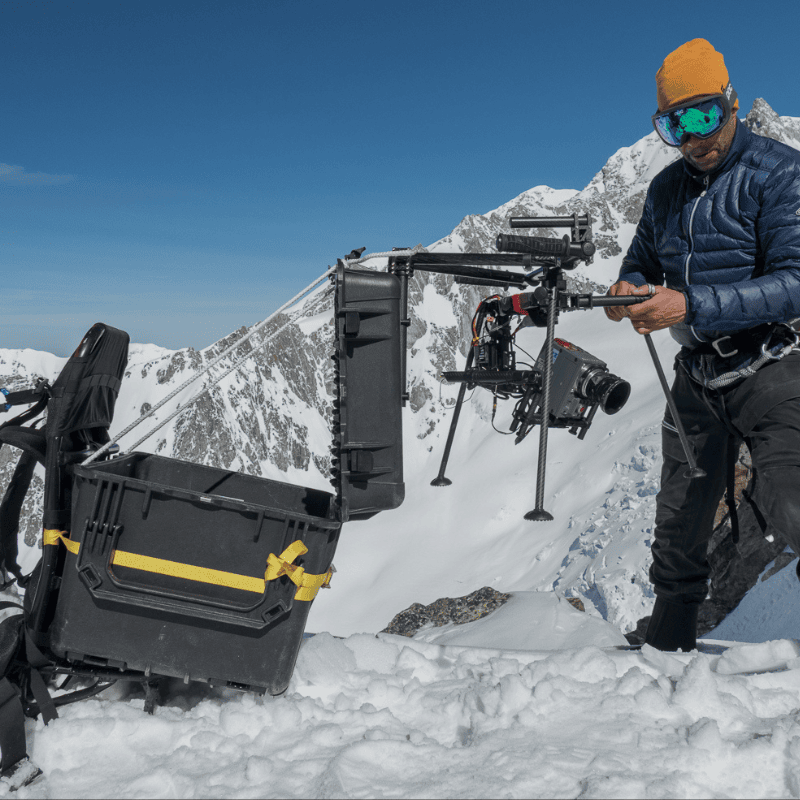 Filmmaker prepares heavy-lift drone with cinema camera on snowy mountain peak for aerial filming in extreme alpine conditions.
